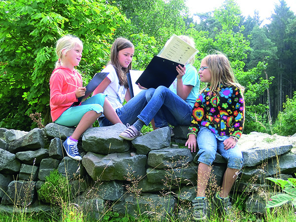 Kinder sitzen auf einer Mauer in der Natur und füllen Fragebögen aus